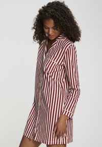 Sieviete ar cirtaini matiem, vilkusi sarkanu un baltu vertikāli svītrainu ilgu krekla kleitu, skatās lejup ar nedaudz smaidu.