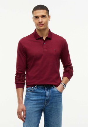 Uomo che indossa una polo bordeaux a maniche lunghe e jeans blu, con una mano in tasca, in piedi davanti a uno sfondo chiaro e uniforme.