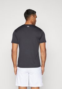 Svart atletisk t-shirt med korta ärmar, har en rund halsringning och en framträdande logodetalj i vitt på baksidan. Slät textur.