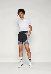 Kvinne står foran en ensfarget bakgrunn iført hvit kortermet Adidas t-skjorte, svart og grå Adidas sykkelshorts, hvite stripete sokker og hvite sneakers.