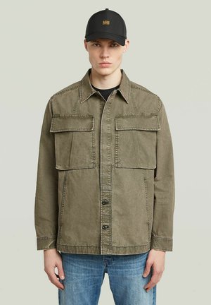 Junger Mann trägt eine verblasste olivgrüne Jacke mit großen Fronttaschen, blaue Jeans und eine schwarze Baseballkappe, steht vor einem einfarbigen Hintergrund.