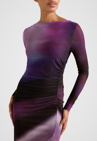 Γυναίκα που φοράει εφαρμοστό, μακρυμάνικο φόρεμα με βαθμιαία μετάβαση χρωμάτων από μοβ σε μαύρο, με ρυθμιζόμενη πτυχώση στο πλάι και λαιμόκοψη τύπου βάρκα.