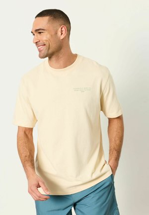 Smiling man with short hair wearing a light beige t-shirt with text and blue shorts, standing with one hand in pocket, looking left.