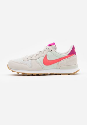 Jasnoszare zamszowe i materiałowe sneakersy z jaskraworóżowym logo Nike, różową zakładką na pięcie, wyściełanym kołnierzem i gumową podeszwą.