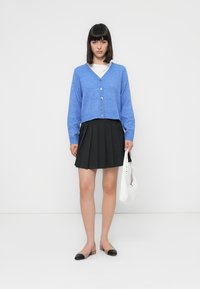 Blauer gestrickter Cardigan mit V-Ausschnitt, fünf Knöpfen, über einem weißen Hemd getragen. Kombiniert mit einem schwarzen Faltenrock und Ballerinas. Weiße Handtasche.