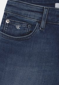 Jeans de denim azul escuro com um bolso frontal apresentando um botão prateado e o logótipo "CK" bordado. O tecido tem uma aparência suave e texturizada.