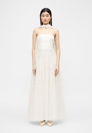 Egyenes fekete hajú nő, fehér, virágos csipkés, pánt nélküli ruhát visel áttetsző réteggel és bézs hegyes orrú cipővel, fehér háttér előtt állva.