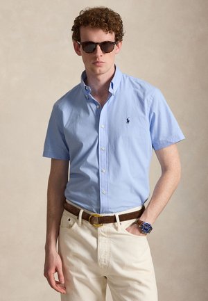 Ung mand iført brune solbriller, lyseblå kortærmet skjorte med knapper, beige bukser, brun bælte og blå ur, poserer mod neutral baggrund.