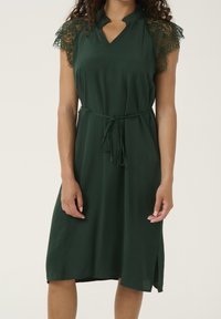 Tmavě zelené šaty s krajkovými krátkými rukávy, výstřihem s průstřihem a páskem v pase, s volným střihem a rozparky na bocích u lemu.