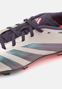 Detailný záber béžovej a čiernej kopačky Adidas na futbal s ružovým logom, čiernymi šnúrkami a iridescentným modrým pruhovaným dizajnom na boku.