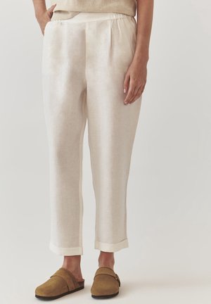 Hellbeige Leinenhose mit umgeschlagenen Säumen, getragen mit braunen Slippern, von der Taille bis zu den Füßen vor einfarbigem Hintergrund gezeigt.