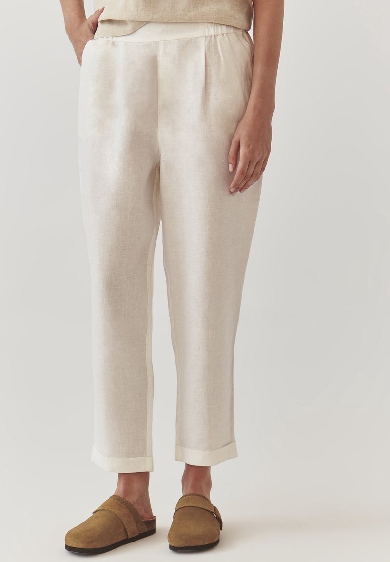 Pantalon en lin beige clair avec ourlets retroussés porté avec des chaussures marron à enfiler, présenté de la taille aux pieds sur fond uni.