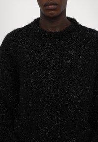 Pulover negru dintr-un material pufos cu o suprafață texturată, având guler rotund și o croială ușor lejeră. Design minimalist, fără modele.