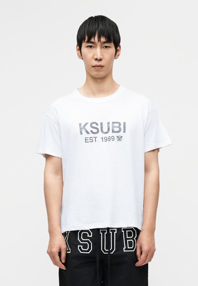 T-shirt en coton blanc à manches courtes, avec un texte imprimé en noir "KSUBI EST. 1999" et un petit logo. Design simple avec une coupe décontractée.