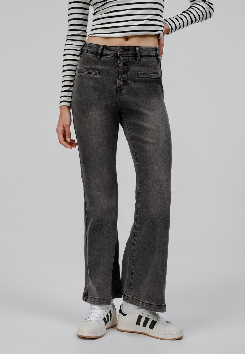 Flared gray denim jeans with a high waist, featuring a button-up front and side pockets. Paired with white sneakers and a black-striped top.