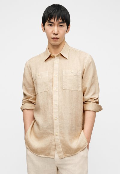 Μπλούζα από μπεζ λινό με κουμπιά μπροστά, δύο τσέπες στο στήθος, μανίκια που έχουν γυριστεί και άνετη εφαρμογή. Λείο ύφασμα με διακριτική γυαλάδα.