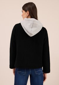 Kvinna som står med ryggen mot kameran, iklädd en svart fleecejacka med en ljusgrå huva och blå jeans mot en beige bakgrund.
