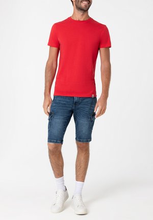 Red cotton t-shirt paired with blue denim knee-length cargo shorts, featuring side pockets. White sneakers and crew-length socks complete the look.