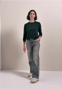 Grünes Langarmshirt, graue verwaschene Jeans, weiße Sneaker mit blauen Akzenten. Das Model steht mit den Händen in den Taschen vor einem neutralen Hintergrund.