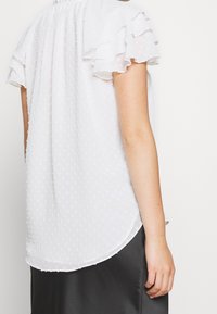 Kvinna i en vit, texturerad blus med korta ärmar och volanger, tillsammans med en svart satängkjol, visad bakifrån mot en vit bakgrund.