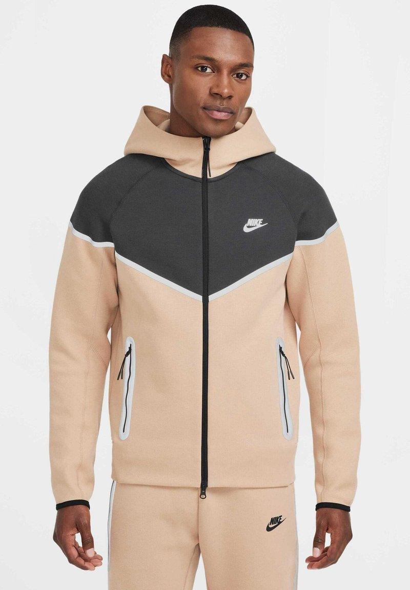 Giacca con cappuccio Nike in beige e grigio scuro, caratterizzata da un design a chevron, cerniera nera e dettagli bianchi. Tessuto liscio, due tasche laterali.