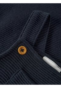 Gros plan sur un tissu en maille côtelée bleu marine avec une bride fixée par un bouton rond beige et une étiquette blanche cousue sur le côté.