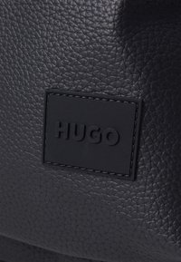 Bolso de cuero negro con una superficie texturizada, logo "HUGO" en relieve en un parche rectangular negro y costuras visibles.