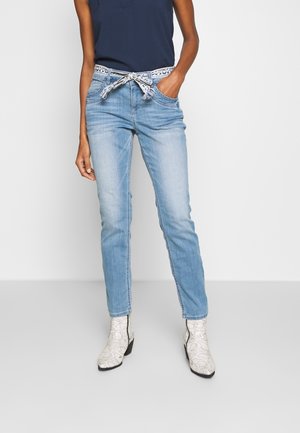 Femme portant un jean bleu clair avec une ceinture blanche à motifs, un haut sans manches bleu marine et des bottines blanches à motif peau de serpent.