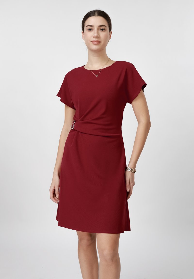 Mujer vistiendo un vestido burdeos hasta la rodilla con mangas cortas y una hebilla lateral, de pie frente a un fondo liso gris claro.