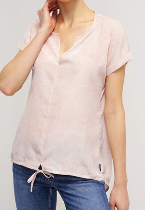Mujer con una blusa de manga corta de color rosa claro con un lazo en el dobladillo y vaqueros azules, mano cerca del cuello.