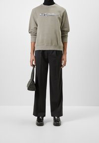 Zelená mikina se stříbrným logem, černé kalhoty a masivní černé boty. Drží malou tmavou tašku; uvolněný střih, minimalistický design.