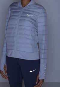 Kobieta w jasnoszarej kurtce puchowej i ciemnych legginsach, obie z widocznymi odblaskowymi logo Nike w kształcie znaku Swoosh.