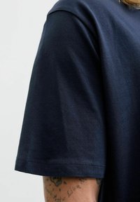 Tamsiai mėlyna medvilninė marškinėliai su apvalia iškirpte ir trumpomis rankovėmis. Lygi tekstūra su subtiliu blizgesiu ir tvarkingu apdailos užbaigimu.