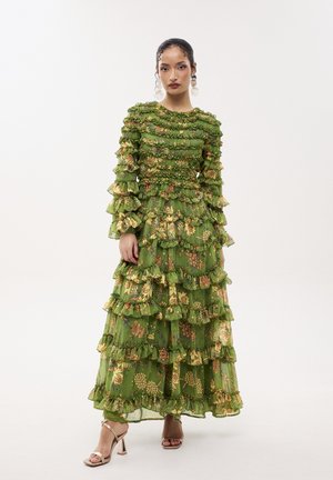 Femme portant une longue robe verte à fleurs avec plusieurs couches à volants et manches longues, debout devant un fond blanc uni.