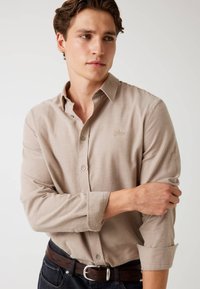 Beige overhemd met lange mouwen en knopen, voorzien van een kraag, een gestructureerde stof en een klein geborduurd logo op de borst. Gecombineerd met een donkere riem.