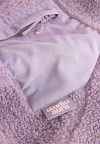 Purpurinė, pūkuota vilnos audinys su lygiu satino pamušalu, turintis raištelį ir etiketę, kurioje parašyta „studio aura“.