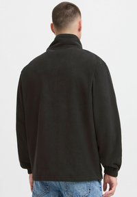 Svart fleecejakke med høykantet krage, lange ermer, elastiske mansjetter og en avslappet passform. Glatt tekstur og minimale designelementer.