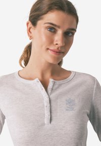Lichtgrijs geribd henley shirt met een ronde halslijn, vijf knopen en een klein geborduurd logo op de linkerborst. Zachte textuur.