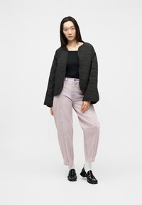 Γυναίκα με μαύρο φουσκωτό μπουφάν, μαύρο τοπ, ανοιχτό ροζ τζιν ψηλόμεσο, λευκές κάλτσες και μαύρα γυαλιστερά μοκασίνια στέκεται μπροστά σε απλό φόντο.