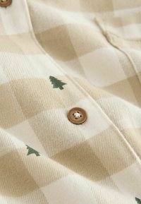 Béžová kostkovaná flanelová košile se zelenou výšivkou stromu a hnědým knoflíkem. Má měkkou texturu a kostkovaný vzor.