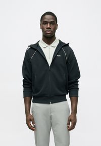 Μαύρο hoodie με φερμουάρ και λευκές γραμμές τόνου, με το λογότυπο "BOSS" στο στήθος, συνδυασμένο με ανοιχτό γκρι πόλο. Ομαλή υφή υφάσματος.