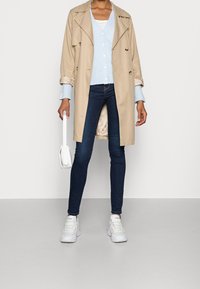 Persona vestida con un abrigo trench beige, un cárdigan de botones azul claro, jeans ajustados oscuros, zapatillas blancas y sosteniendo un pequeño bolso blanco.