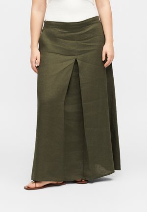 Mujer con una falda larga color verde oliva con abertura frontal, top blanco y sandalias planas marrones sobre un fondo liso.