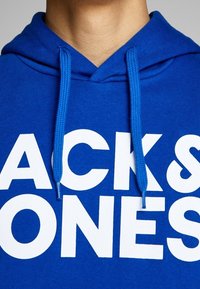 Ingrandimento di una persona che indossa una felpa blu con cappuccio, con spesse corde di regolazione e grande testo bianco con la scritta "JACK & JONES" sul petto.
