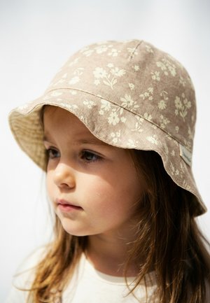 Junges Kind mit langen braunen Haaren, das einen beigen Anglerhut mit einem weißen Blumenmuster trägt und leicht zur Seite schaut.