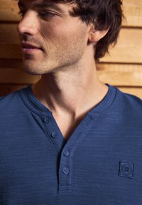 Man met kort bruin haar, gekleed in een blauw henley shirt met vier knopen, kijkt naar links tegen een houten muurachtergrond.