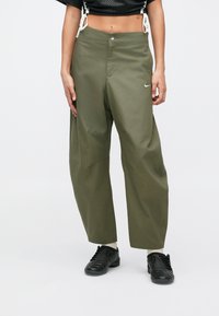 Olivgröna byxor med vida ben, framknäppning, sidfickor och en liten vit logga på höger sida. Mjuk tygkänsla.