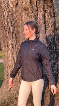 Jeune femme portant une veste noire et des leggings beiges, debout les yeux fermés, souriante, devant un grand tronc d'arbre texturé au soleil.