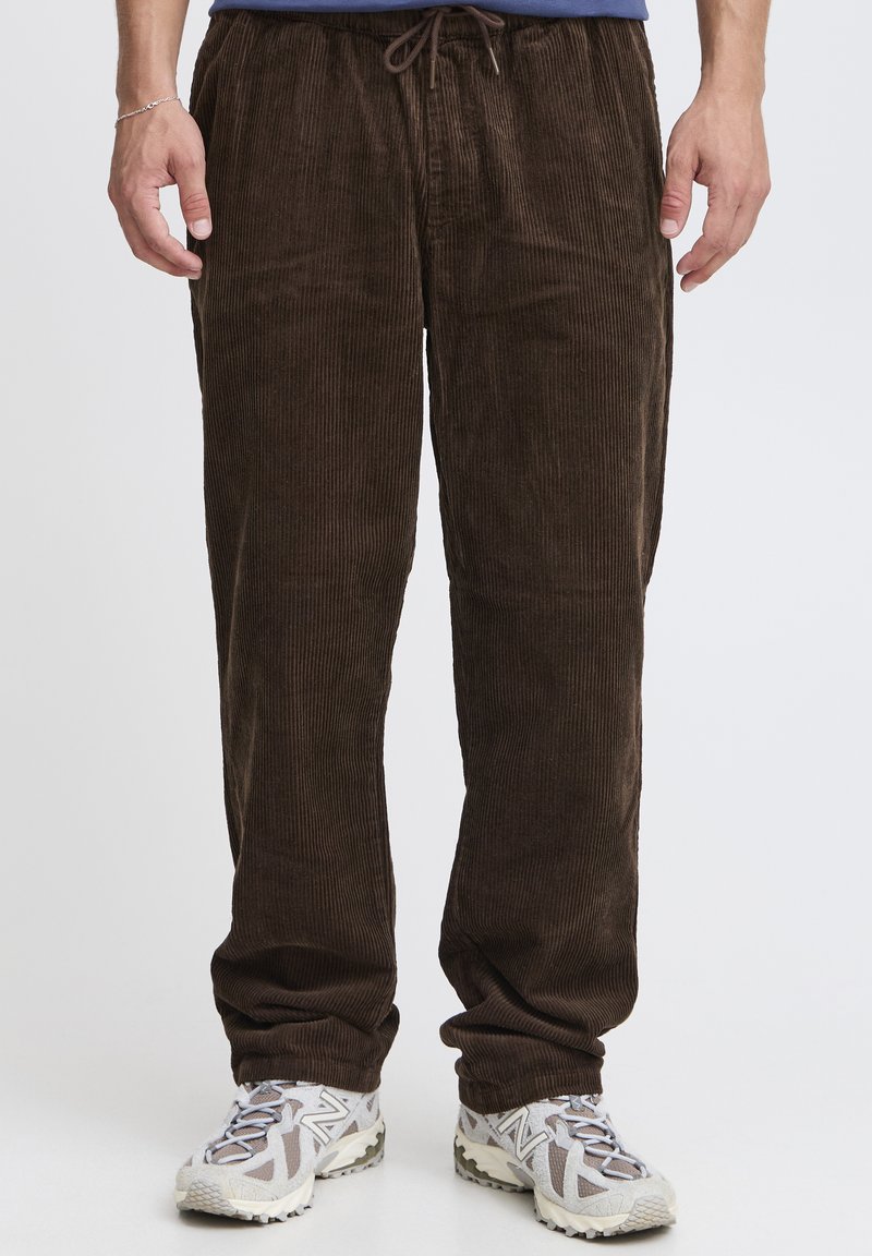 Pantaloni marroni in velluto a coste con trama verticale a coste, vita con coulisse e vestibilità comoda, abbinati a sneakers grigio chiaro.