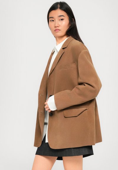 Donna con lunghi capelli scuri che indossa una camicia bianca, una giacca oversized marrone e una gonna scura, in posa contro uno sfondo grigio chiaro uniforme.